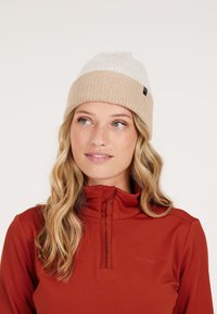 Beige en witte gebreide beanie met ribbelstructuur, voorzien van een klein zwart merklabel. G gedragen met een rode top, die een hoge kraagontwerp heeft.