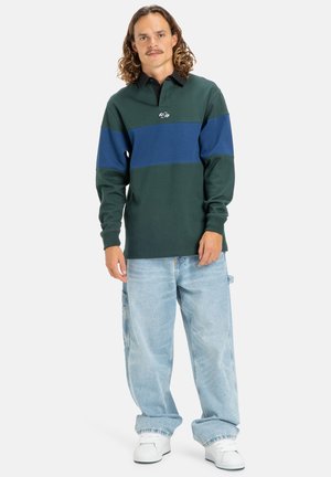 Grön- och blårandig långärmad polo tröja med svart krage, tillsammans med ljusblå oversized jeans och vita sneakers.