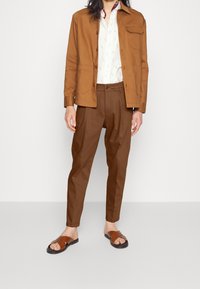 Brązowa kurtka na guziki noszona na białej koszuli z subtelnym wzorem. Brązowe spodnie i brązowe sandały dopełniają strój. Prosty, casualowy design.