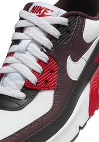 Nike tenisice imaju gornji dio od bijele i crne kože s burgundskim i crvenim akcentima. Debele bijele vezice i teksturirani bočni paneli upotpunjuju dizajn.