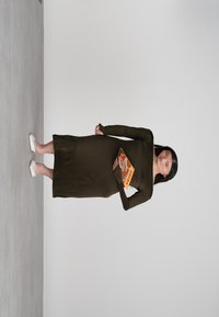 Frau in einem langen olivgrünen Kleid und weißen Schuhen, die ein oranges Buch mit dem Titel "Chika" vor einem schlichten hellgrauen Hintergrund hält.