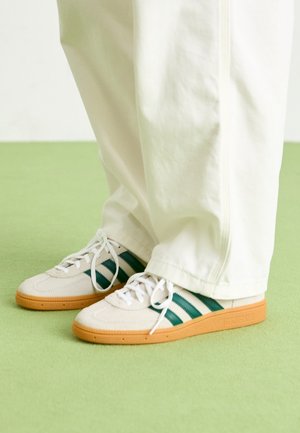 Personne portant un pantalon blanc et des baskets beiges avec des bandes vert foncé et des semelles en gomme, debout sur un sol vert clair.
