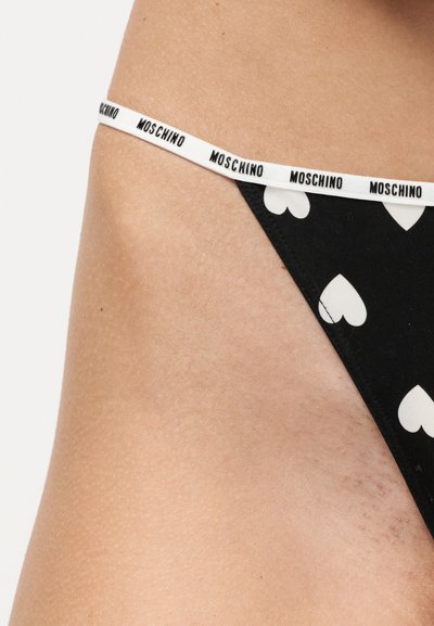 Κοντινό πλάνο μαύρου εσωρούχου με λευκά μοτίβα καρδιάς και λευκή ελαστική ζώνη με επαναλαμβανόμενο κείμενο «Moschino» στη γοφά ενός ατόμου.