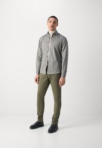 Camisa de botones en gris con pequeños lunares negros, combinada con pantalones ajustados verde oliva y mocasines negros, sobre un fondo blanco.