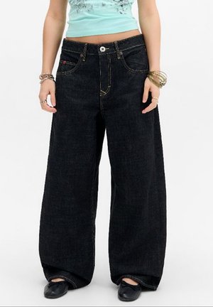 Person, die weite schwarze Jeans, schwarze flache Schuhe, ein hellblaues, kurzes Oberteil und mehrere Armbänder an beiden Handgelenken trägt.