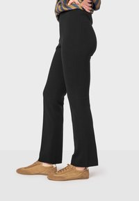 Pantalons évasés noirs en tissu doux, dotés d'une taille haute. Associés à des baskets en suède marron clair, présentant une texture lisse.