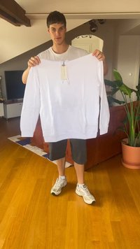 Giovane uomo in piedi all'interno che tiene una felpa bianca a maniche lunghe con un'etichetta davanti a un divano marrone e una pianta in vaso.