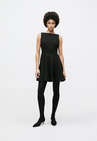 Μαύρο φόρεμα χωρίς μανίκια με εφαρμοστή μπούστα και φουσκωτή φούστα. Λείο ύφασμα, μήκος μέχρι το γόνατο, συνδυασμένο με μαύρες κάλτσες και μυτερές γόβες.