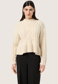 Creme kabelstrik sweater med høj hals, ribbet kant og struktureret overflade, parret med sorte skræddersyede bukser.