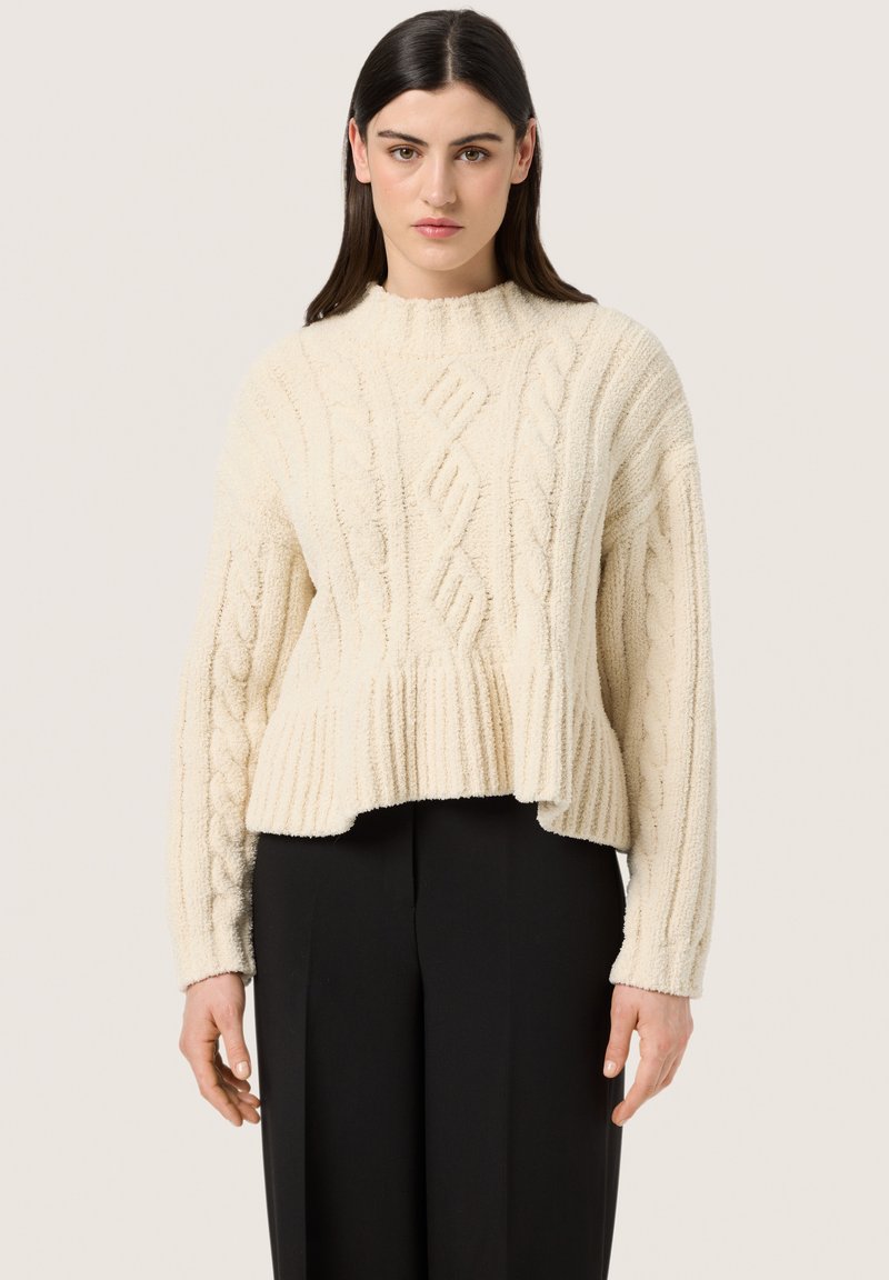 Creme kabelstrik sweater med høj hals, ribbet kant og struktureret overflade, parret med sorte skræddersyede bukser.