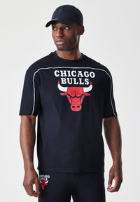 Črna majica s kratkimi rokavi z rdečim logotipom Chicago Bulls in belimi detajli. Nosena z črnimi hlačami, ki prikazujejo sproščen športni slog.