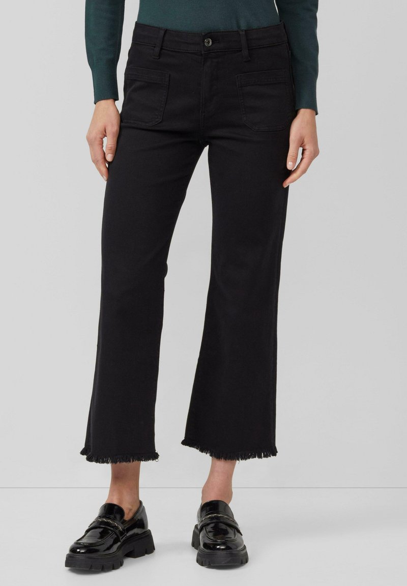 Femme portant un pantalon flare noir court avec des ourlets effilochés et des mocassins en cuir verni noirs, se tenant contre un fond clair.