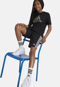 Camiseta negra de manga corta con logo de estampado de leopardo, combinada con pantalones cortos negros con rayas blancas. Zapatillas blancas y calcetines a rayas.