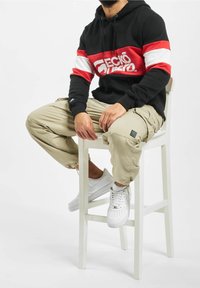Sudadera negra con rayas horizontales rojas y blancas y logo. Pantalones cargo beige con bolsillos. Zapatillas blancas con perforaciones.