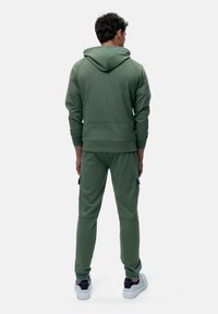 Grüne Baumwollkapuzenjacke mit einem strukturierten Design auf der Rückseite, kombiniert mit passenden schmalen Hosen und weißen Sneakers, von hinten betrachtet.