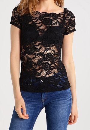 Vrouw draagt een zwart kort mouwbloesje van bloemenkant over een zwarte beha, gecombineerd met blauwe spijkerbroeken, tegen een effen achtergrond.