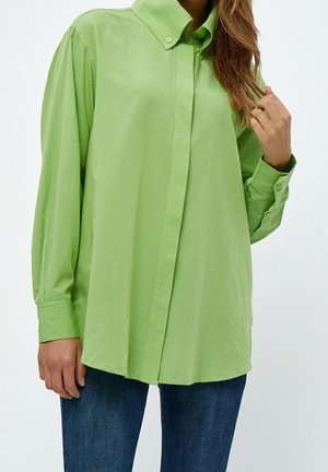 Chemise verte à col, fabriquée en tissu léger, avec des manches longues, une coupe décontractée et des boutons sur le devant. Portée avec un jean noir.
