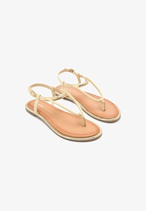 Sandalias beige con diseño de tira en T, material suave y suela acolchada en tono tostado. Presentan una delgada correa para el tobillo con hebilla y detalles metálicos.