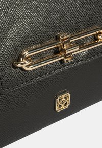 Close-up van een zwarte gestructureerde leren handtas met een gouden rechthoekige sluiting en een klein decoratief gouden embleem eronder.
