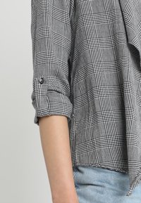 Košile s černobílým kostkovaným vzorem, s ohrnutými rukávy, knoflíkovými detaily a měkkou látkou. Spodní okraj je mírně asymetrický.