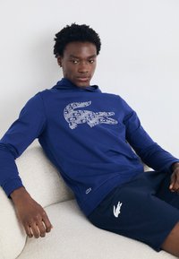 Marineblauer Hoodie mit einem grafischen Krokodil-Design in Weiß. Hergestellt aus weichem Material mit lockerer Passform. Kombiniert mit passenden Shorts, die ein Logo tragen.