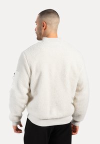 Hombre con cabello corto que lleva una chaqueta de forro polar blanca texturizada y pantalones negros, mirando hacia atrás, frente a un fondo liso.