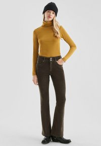 Junge Frau in senfgelben Rollkragenpullover, dunkelbraunen Cordhosen, schwarzem Beanie und schwarzen Stiefeln, die vor einem schlichten Hintergrund steht.