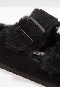 Sandali in suede neri con dettagli in pelliccia e chiusura a doppia fibbia. La superficie texturizzata e la suola sagomata offrono comfort e stabilità.