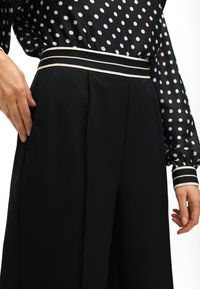 Black wide-leg trousers featuring a striped waistband. The fabric is smooth with a matte finish, complementing a polka-dot blouse.