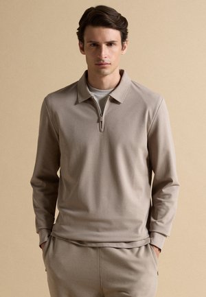 Giovane uomo che indossa un top beige con maniche lunghe e cerniera e pantaloni abbinati, in piedi con le mani nelle tasche davanti a uno sfondo beige uniforme.