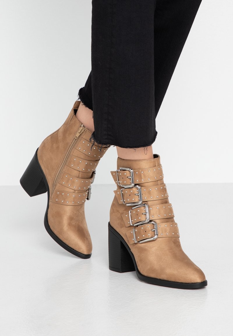 Bottes chevilles en suède beige avec un large talon noir, dotées de trois sangles à boucles ornées de clous en argent et d'une fermeture éclair sur le côté.