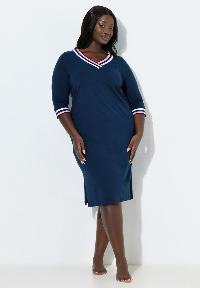 Frau steht barfuß, trägt ein marineblaues knielanges Kleid mit dreiviertel Ärmeln und pink-weiß gestreiften Abschlüssen an Ausschnitt und Bündchen.