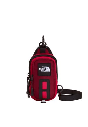 Mała czerwona saszetka North Face z czarnym zamkiem, naszywką z logo, różowym detalem paska i odpinanym czarnym paskiem na nadgarstek, wisząca na białym tle.