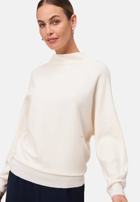 Femme portant un pull en ribbed crème avec des manches longues bouffantes et un col montant, associé à un pantalon sombre, se tenant contre un fond blanc.