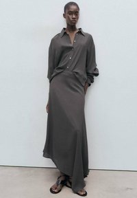 Un vestido largo de color gris oscuro, con un top de camisa abotonada, falda larga y dobladillo asimétrico. Combinado con sandalias negras planas sobre un fondo claro.