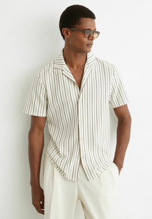 Mees, kes kannab prille ja lühikeste varrukatega valget särki, millel on õhukesed vertikaalsed pruunid triibud, ning heledaid pükse, seisab valge seina taustal.