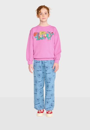 Bambino dai capelli rossi che indossa una felpa rosa con motivi floreali, pantaloni blu larghi con testo stampato e scarpe da ginnastica rosse, in piedi davanti a uno sfondo chiaro.