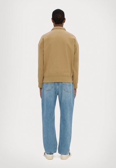 Tan sweatshirt with a relaxed fit, ribbed cuffs, and hem, paired with loose light blue jeans and off-white sneakers.