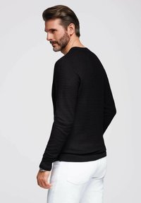 Schwarzer strukturierter Pullover mit rundem Ausschnitt, langen Ärmeln und gerippten Bündchen, kombiniert mit weißen Hosen und von hinten betrachtet.
