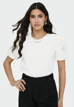 Vrouw met lang donker haar draagt een witte blouse met pofmouwen en kantdetails, gecombineerd met een zwarte broek met hoge taille, poseert met handen in de zij.