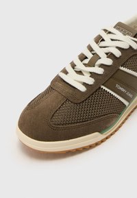 Gros plan sur une basket Tommy Jeans marron avec des lacets blancs, une tige en mesh et en daim, une semelle crème et une semelle extérieure en gomme texturée.