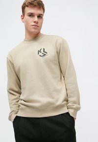 Beige színű pulóver, puha anyagból készült, kerek nyakkivágással és hosszú ujjal rendelkezik. Az elején fekete geometrikus logó található.