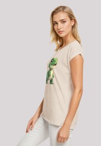 Beige short-sleeve t-shirt featuring a large green dinosaur graphic on the front. The shirt has a relaxed fit and a soft texture.