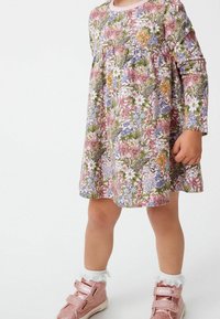 Enfant portant une robe à manches longues à motifs floraux avec des motifs roses, blancs et verts, des chaussettes blanches à volants et des chaussures roses scintillantes.