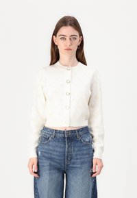 Cream cardigan with a round neck, long sleeves, and five button closures. Features a subtle pattern and cropped design, paired with blue jeans.