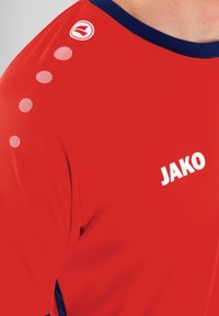 Rotes Sportshirt mit marineblauem Kragen, versehen mit fünf weißen Punktemuster-Akzenten auf der Schulter und einem weißen "JAKO"-Logo auf der Vorderseite.