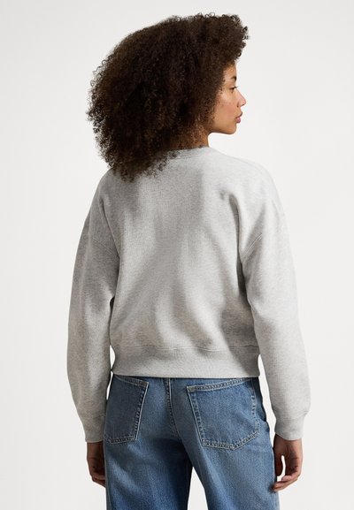 Mulher com cabelo encaracolado, vestindo um casaco cinza claro e jeans azuis, vista de costas contra um fundo branco simples.