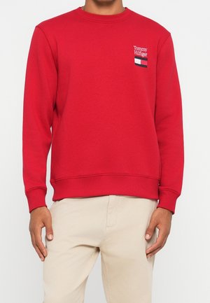 Mand iført rød Tommy Hilfiger sweatshirt med logo på venstre bryst og beige bukser, stående foran en ensfarvet hvid baggrund.