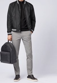 Muž v černé varsity bundě, černém svetru, šedých kalhotách a černých teniskách, držící černý kožený batoh v levé ruce.