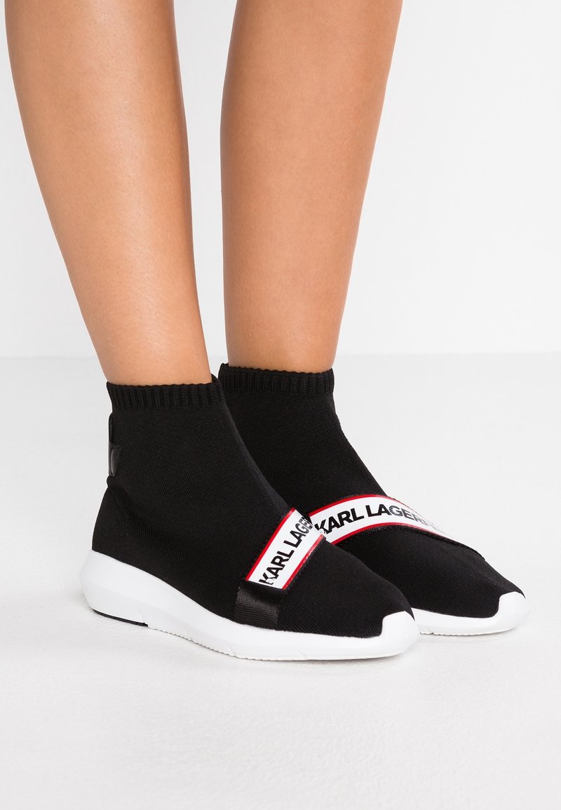 Black sock-style sneakers with a ribbed cuff, white sole, and Karl Lagerfeld branded strap featuring black text on a red background.